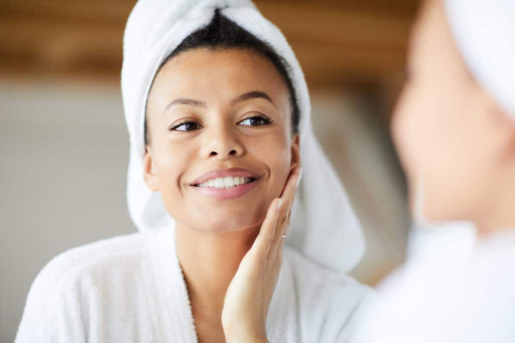 Woman with clear skin smiling in mirror, towel wrapped around hair